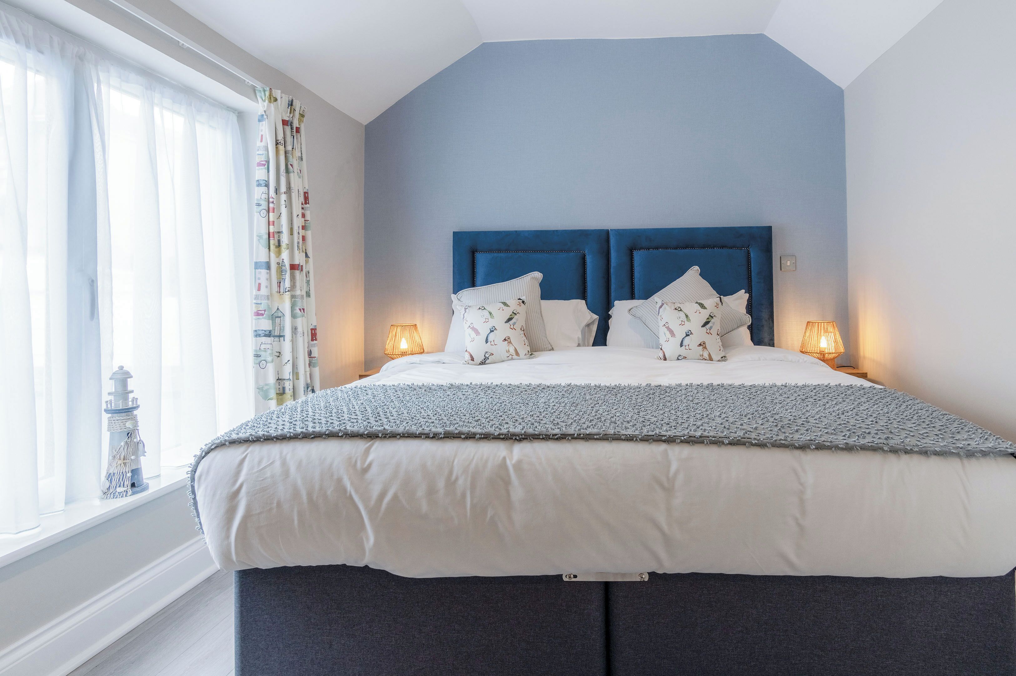 Una cama bien hecha con un cabecero azul, ropa de cama blanca y almohadas estampadas. Hay dos lámparas de noche a cada lado de la cama.