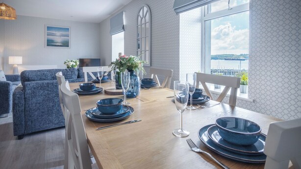 A dining area with a wooden table set for four, blue dinnerware, and white chairs. - Haven View - 2 Bedroom Apartment - Milford Haven (Milford Haven)