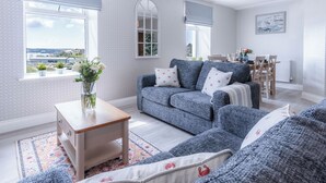 A living room with a grey sofa, a wooden coffee table, and a view of the outdoors through a large window. - Haven View - 2 Bedroom Apartment - Milford Haven (Milford Haven)
