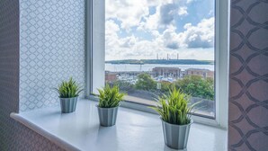 A window with three potted plants on a white ledge, overlooking a marina with boats and buildings. - Haven View - 2 Bedroom Apartment - Milford Haven (Milford Haven)