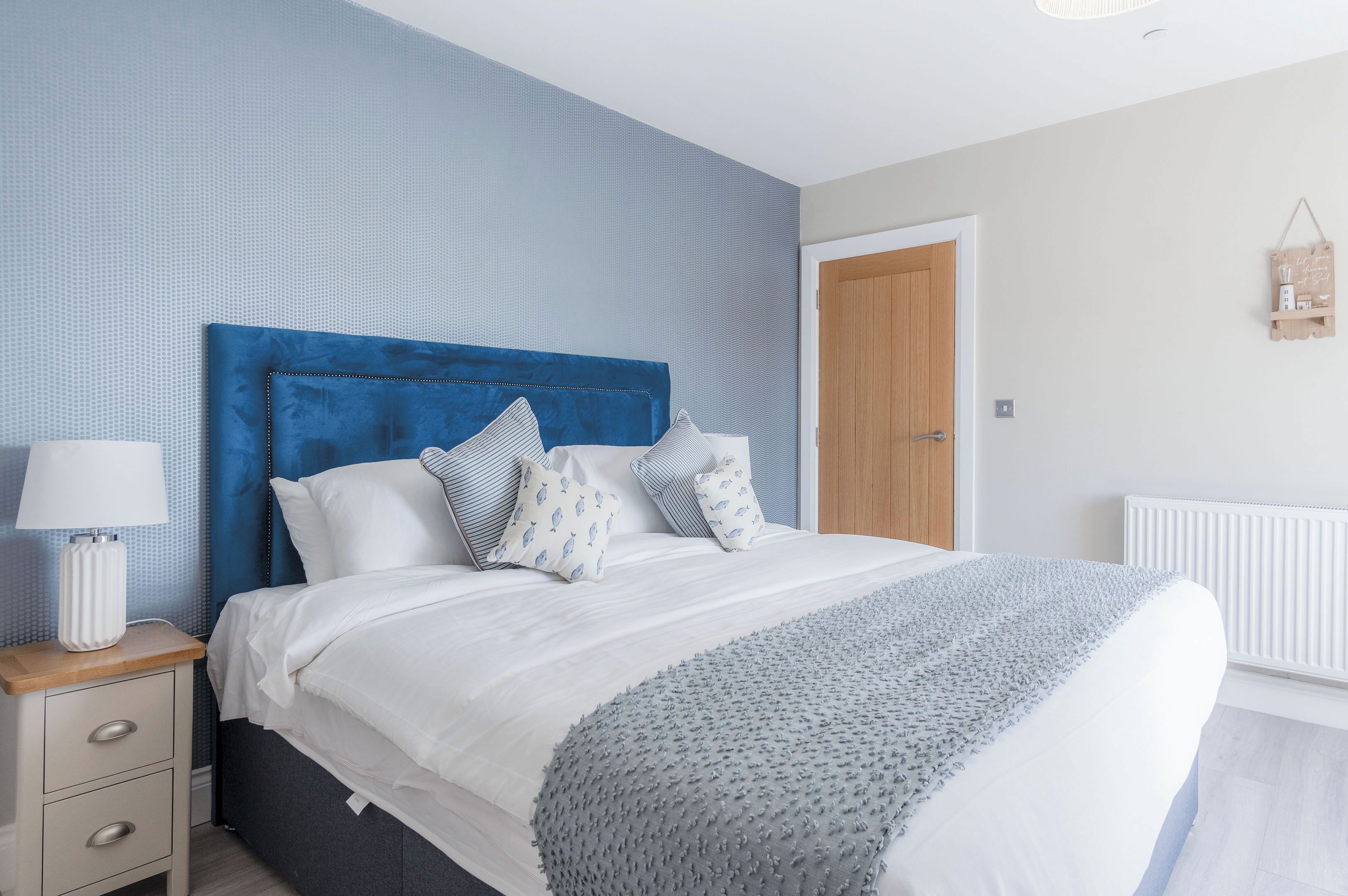 Een keurig opgemaakt bed met een blauw hoofdbord, een nachtkastje met een lamp en een wandplank met een klein voorwerp.