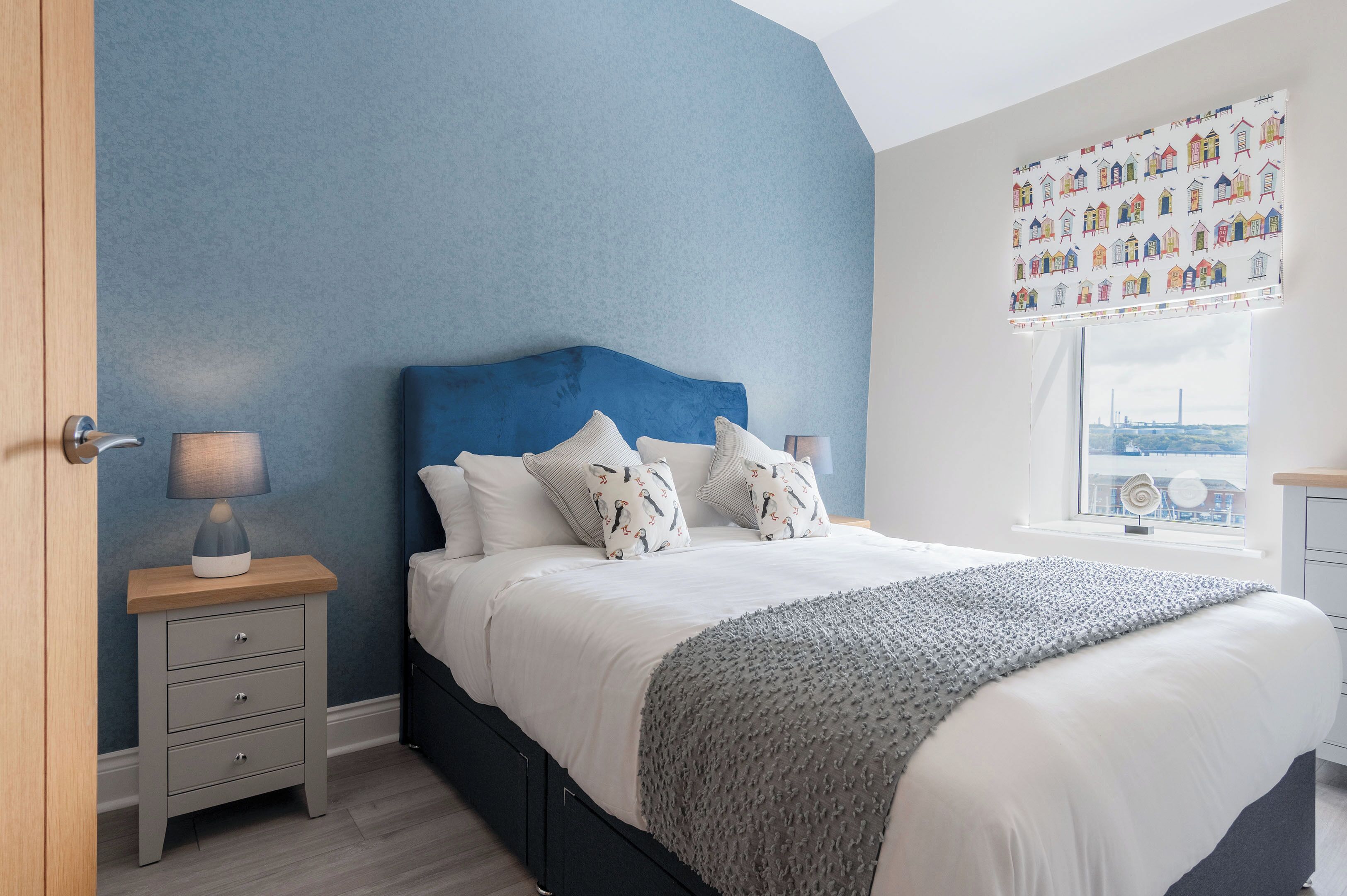 A bedroom with a blue wall, a bed with white bedding, patterned pillows, a grey blanket, a bedside table with a lamp, and a window with a city view.