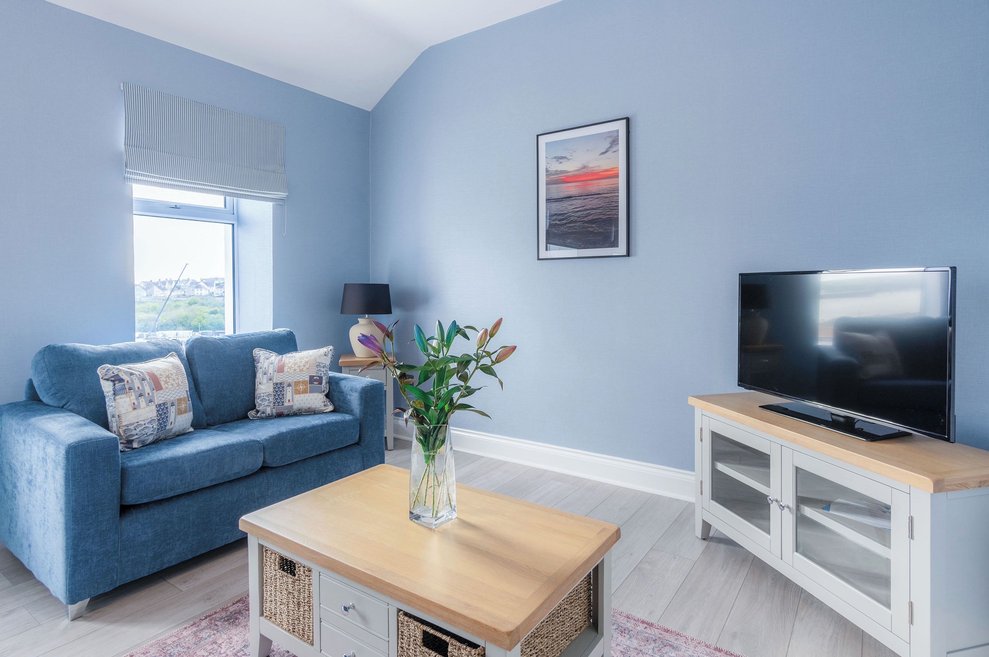 A living room with a blue sofa, a wooden coffee table, a TV on a cabinet, and a window with a view.