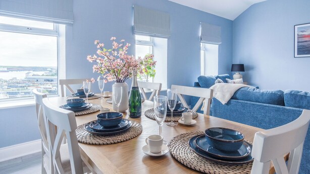 A dining table set with blue plates, white cups, and a vase of pink flowers. - Harbour Heights - 2 Bed Apartment - Milford Haven (Milford Haven)