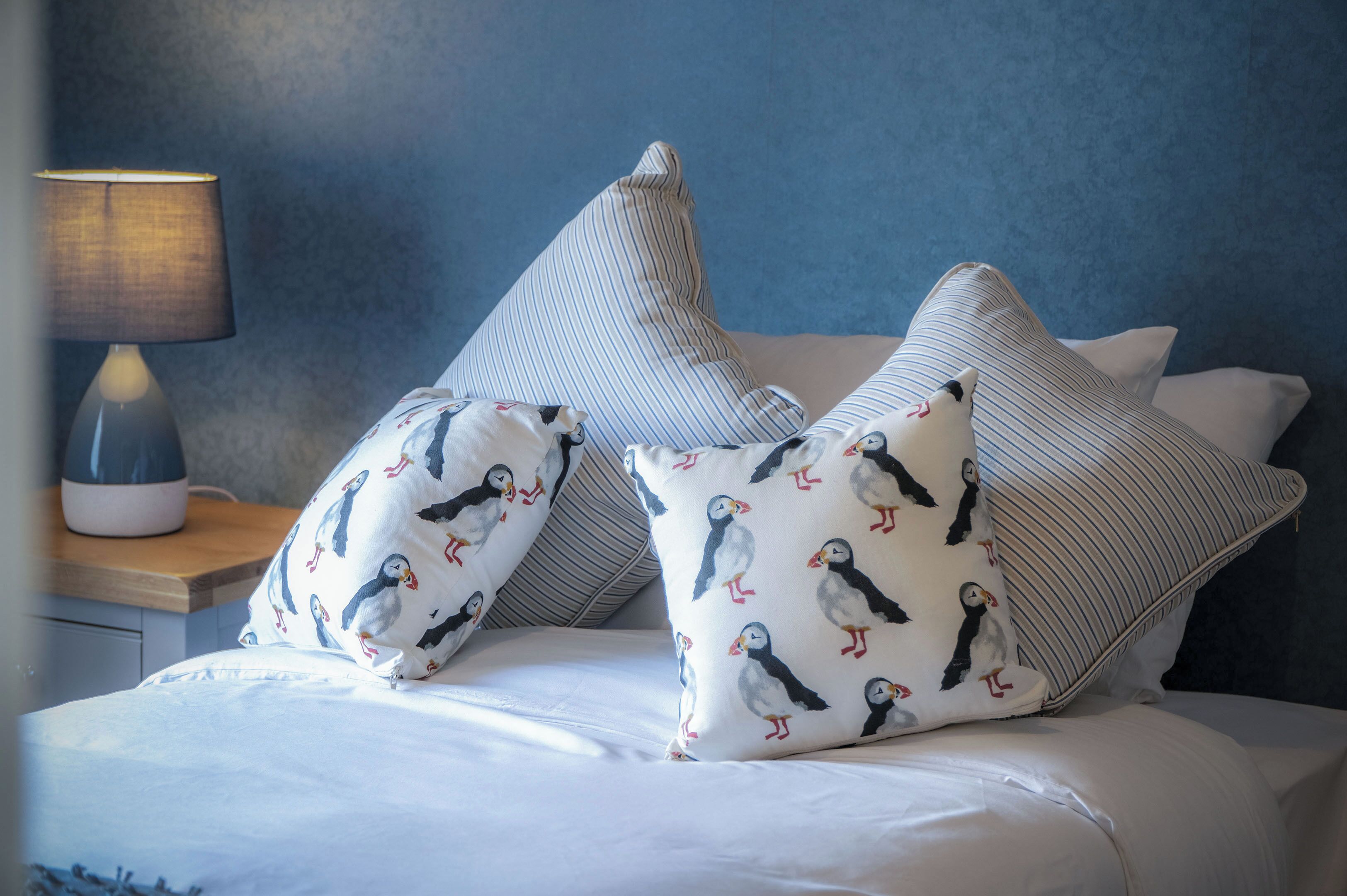 A bed with white bedding and pillows featuring a puffin pattern, a bedside lamp, and a blue textured wall.