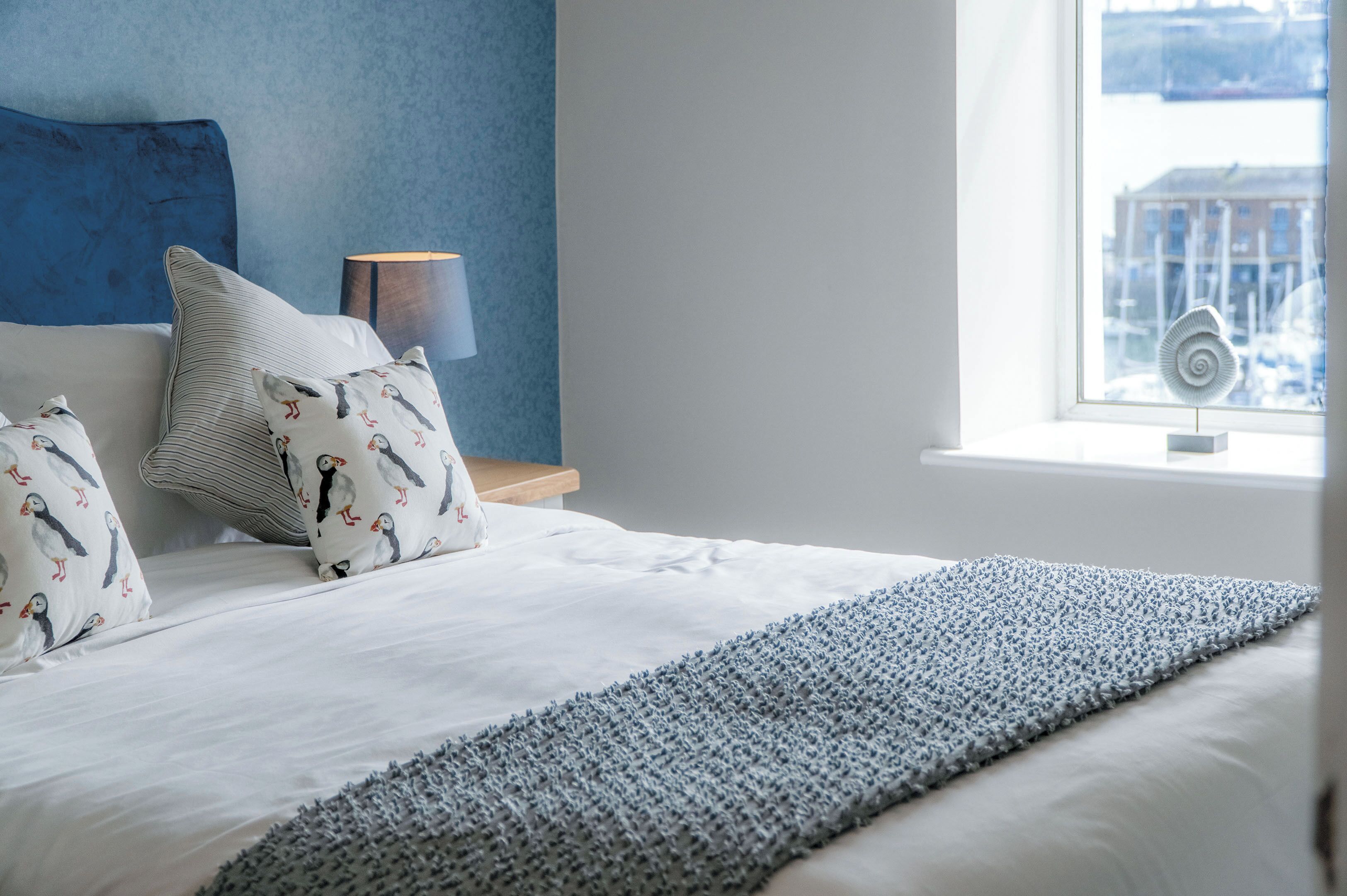 A neatly made bed with a blue headboard, a bedside lamp, and a view of a snowy landscape through a window.