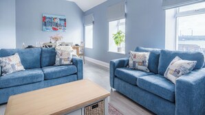 A modern living room with two blue sofas, a wooden coffee table, and a dining area with a table and chairs. - Harbour Heights - 2 Bed Apartment - Milford Haven (Milford Haven)
