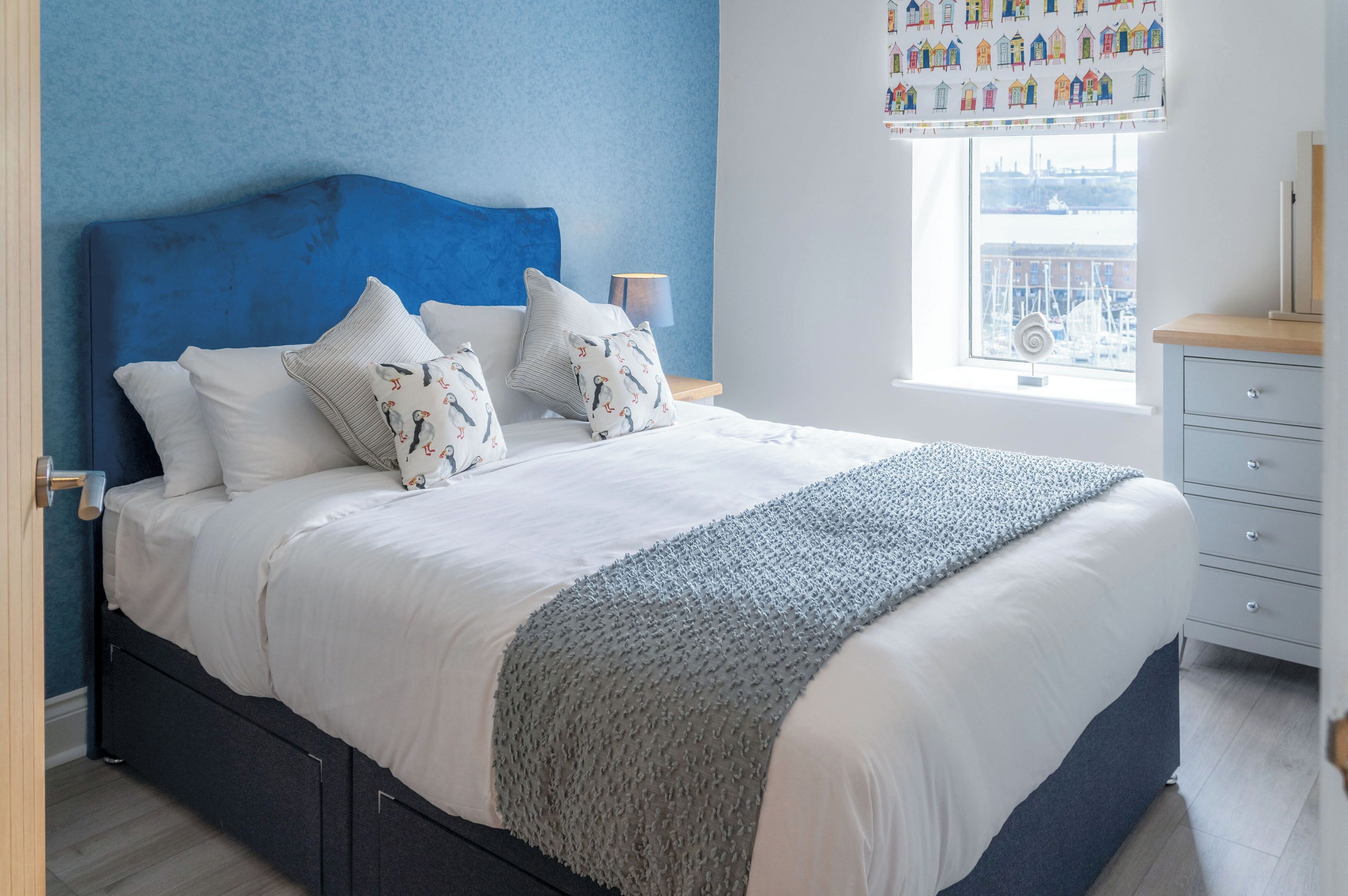 Een net opgemaakt bed met witte lakens en gestreepte kussens, een blauw hoofdeinde en een raam met uitzicht op een steiger.