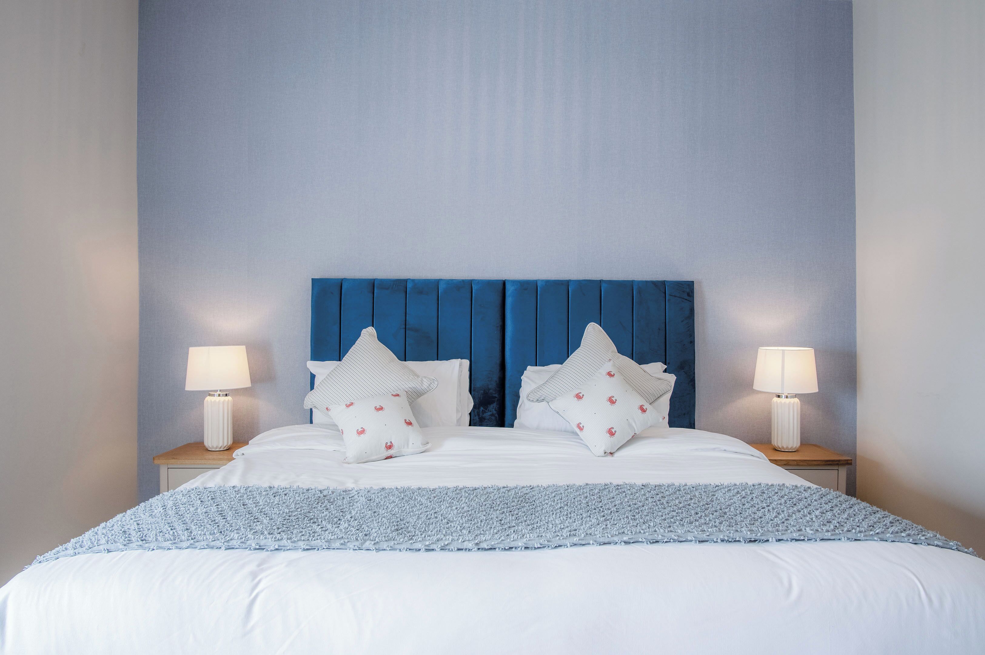 A neatly made bed with white bedding and blue accents, flanked by two bedside tables with lamps, against a blue headboard.
