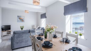 A modern dining area with a wooden table set for four, a grey sofa, and a city view through large windows. - Beach Haven Hideout - 2 Bedroom - Milford Haven (Milford Haven)