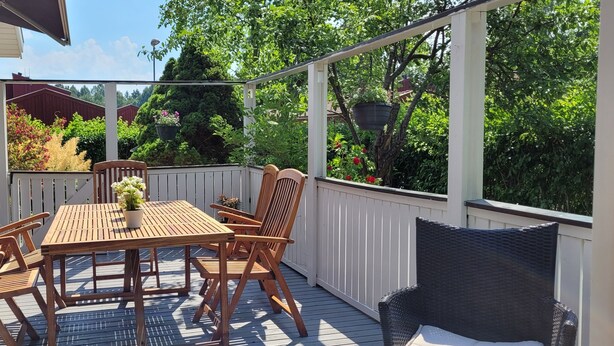 Une terrasse en bois, agrémentée d’une table et de chaises, entourée d’une balustrade blanche, et donnant sur un jardin où poussent différentes plantes et arbres.