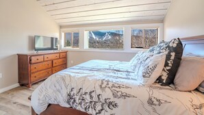 A bedroom with a bed, a dresser, a TV, and a mountain view through the windows. - Loon Mountain View at Ravine (Lincoln)