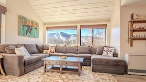A cozy living room with a sectional sofa, a wooden coffee table, and a large window offering a mountain view. - Loon Mountain View at Ravine (Lincoln)