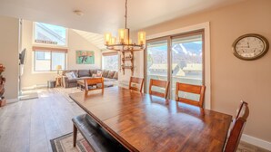 A spacious dining area with a large wooden table, multiple chairs, and a chandelier. There is a sofa and a coffee table in the background, and a large window offering a view of mountains. - Loon Mountain View at Ravine (Lincoln)