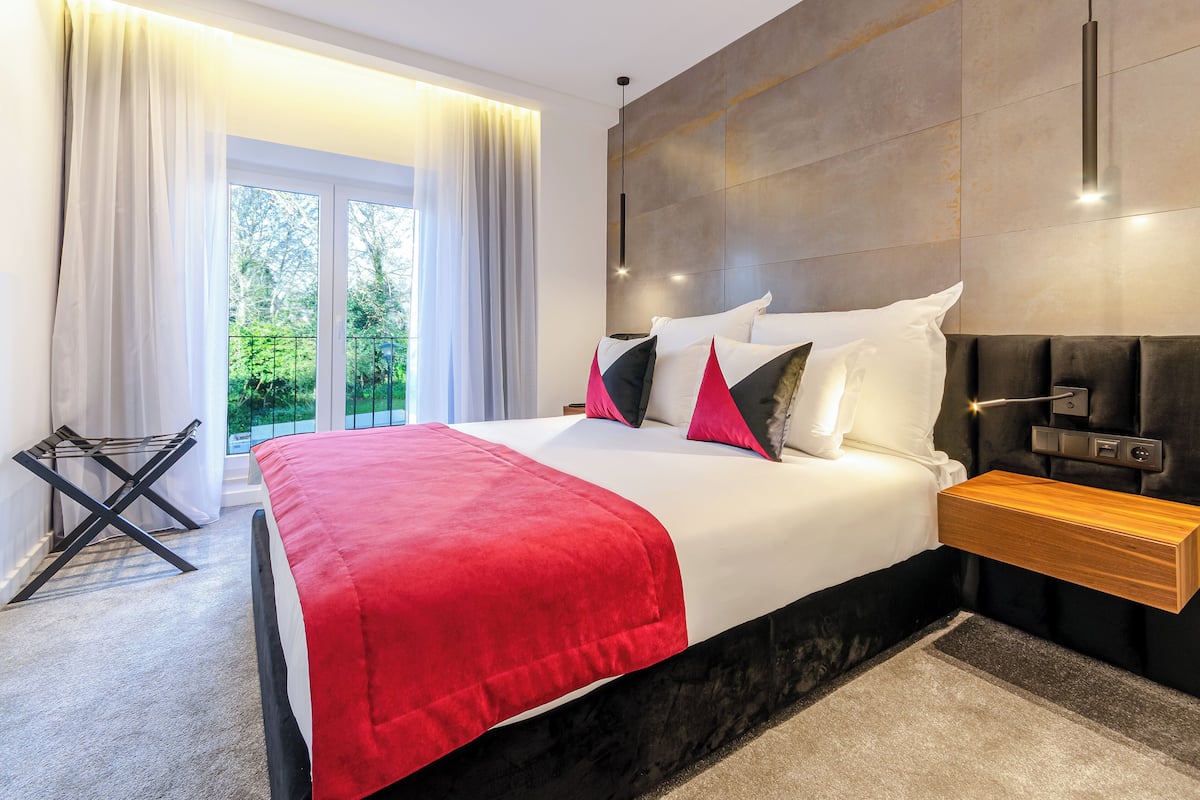 A modern bedroom with a large bed, a wooden bedside table, and a view of greenery through the window.