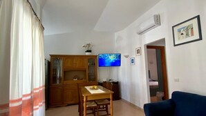 Un salon avec un canapé bleu, un fauteuil rouge, une table à manger en bois avec des chaises, un téléviseur et un coin cuisine équipé de placards et d’un climatiseur.