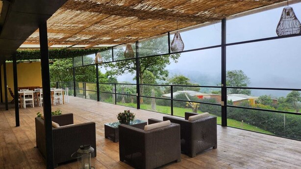 Un vaste espace extérieur aménagé avec des meubles en osier et doté d’un toit de chaume, offrant une vue sur un jardin luxuriant et des bâtiments situés au loin.