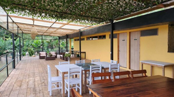 An outdoor patio with wooden flooring, white and wooden tables and chairs, and a yellow wall with a door.