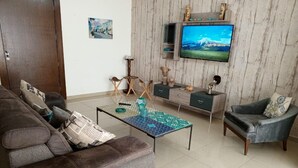A living room with a brown sofa, a patterned coffee table, a grey armchair, and a TV mounted on a wooden wall. - Tilal Fanar Resort, Pools, Generator Included (Ain Saadeh)