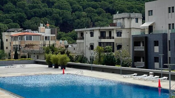 A swimming pool with lounge chairs and a view of buildings and a forested hill. - Tilal Fanar Resort, Pools, Generator Included (Ain Saadeh)