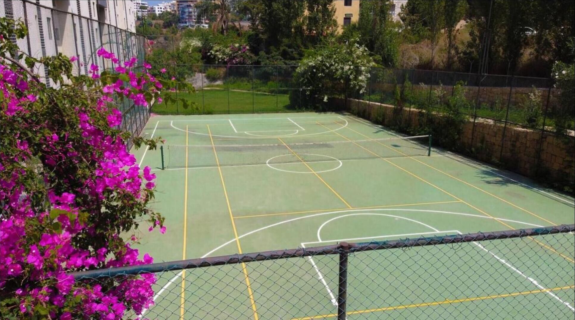 An outdoor basketball court with a chain-link fence, surrounded by greenery and buildings.