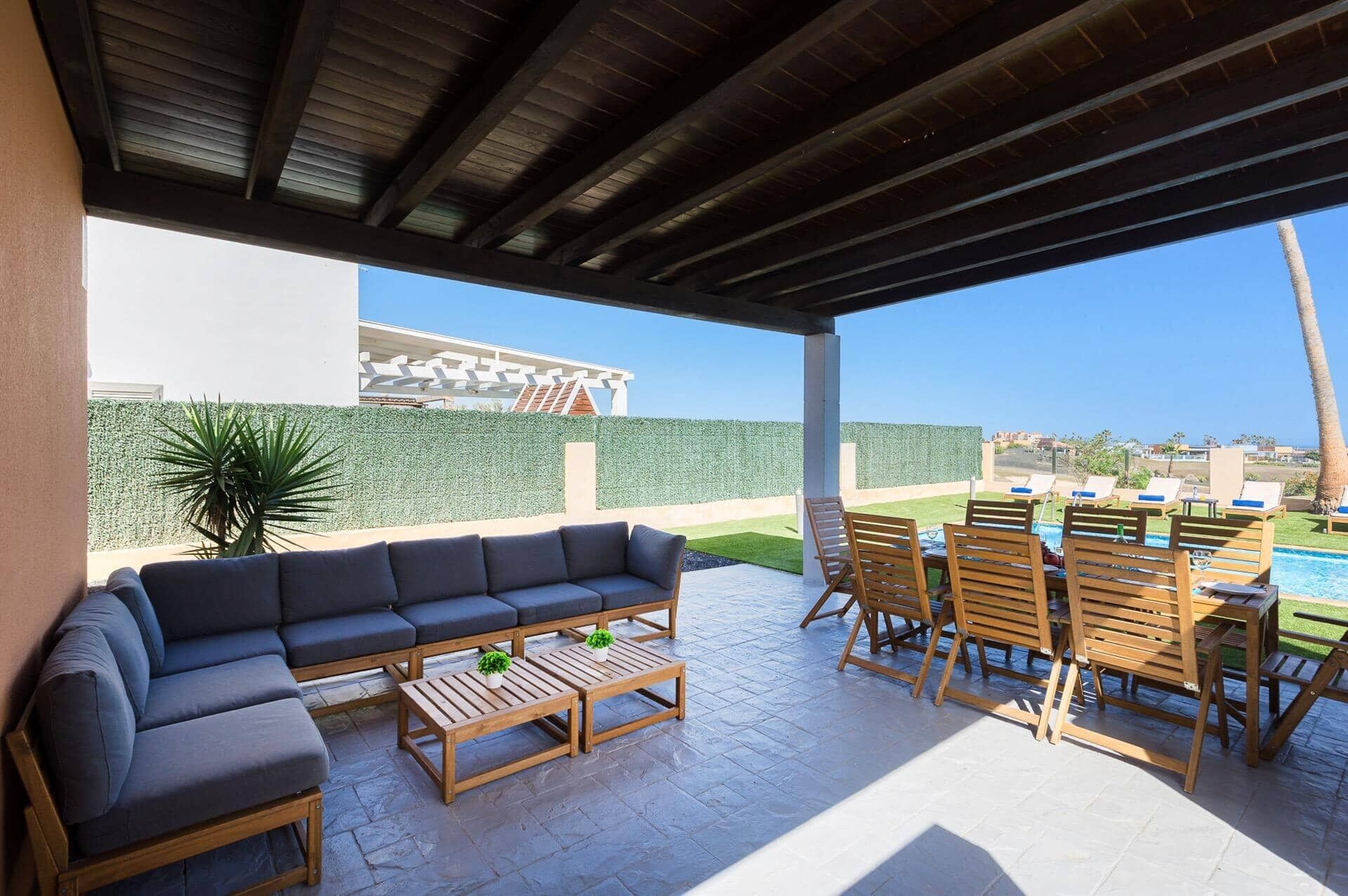 A covered outdoor area with a sofa, wooden tables, and lounge chairs.