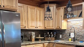 Una cocina con armarios de madera, encimera de granito, refrigerador de acero inoxidable y dos lámparas colgantes.