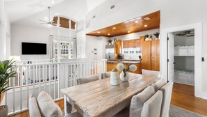 Un'ampia zona giorno con un tavolo da pranzo in legno, arredi bianchi, una TV a parete e un angolo cottura con mobili in legno e una ventola a soffitto.