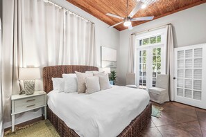 Un dormitorio con una cama grande, una mesita de noche, una lámpara, un ventilador de techo y vistas a través de una puerta.