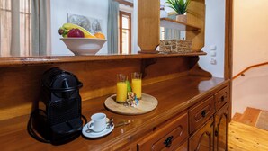 A wooden buffet table with a bowl of fruit, two glasses of orange juice, a coffee maker, and a cup on a saucer.