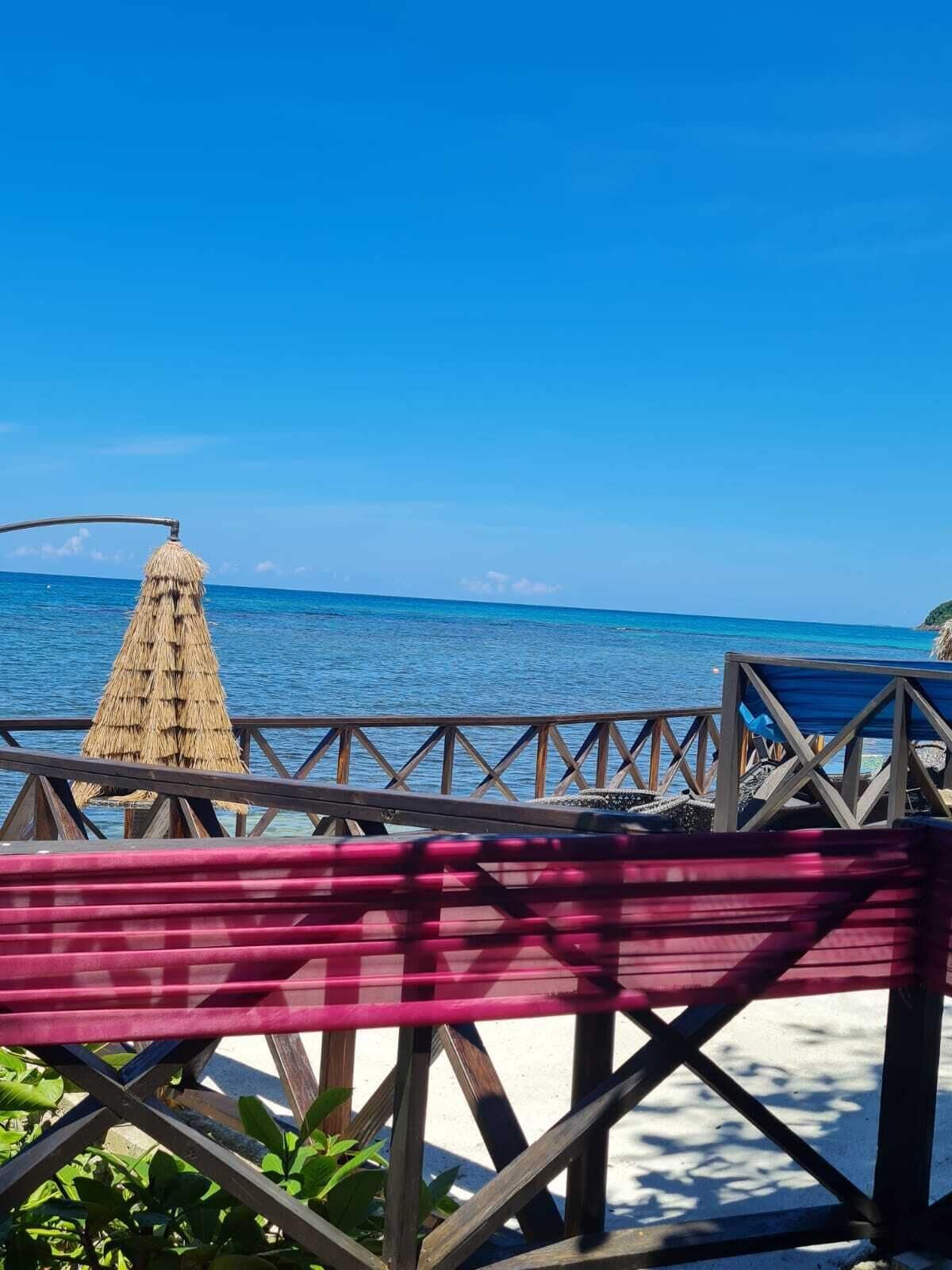 Vista para a praia com um deck de madeira, um guarda-sol de palha e uma rede.