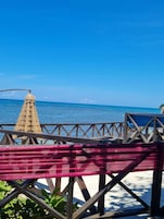 Vista de la playa con una terraza de madera, un paraguas de paja y una hamaca.