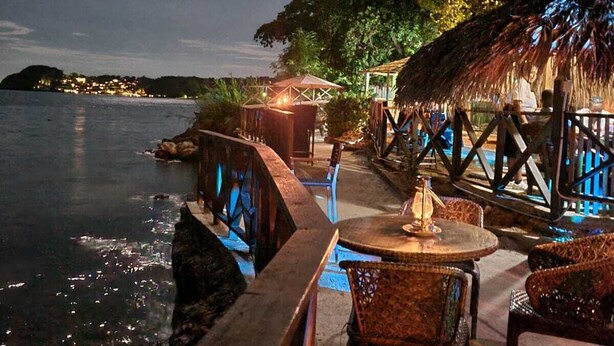 An outdoor dining area with a view of the sea, featuring a thatched-roof structure and wicker furniture. - Sky Beach Rooms (Montego Bay)