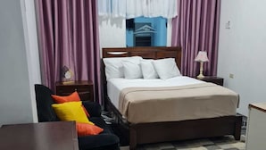 A bedroom with a bed, a chair, a nightstand, a lamp, and a window with curtains. - Sky Beach Rooms (Montego Bay)