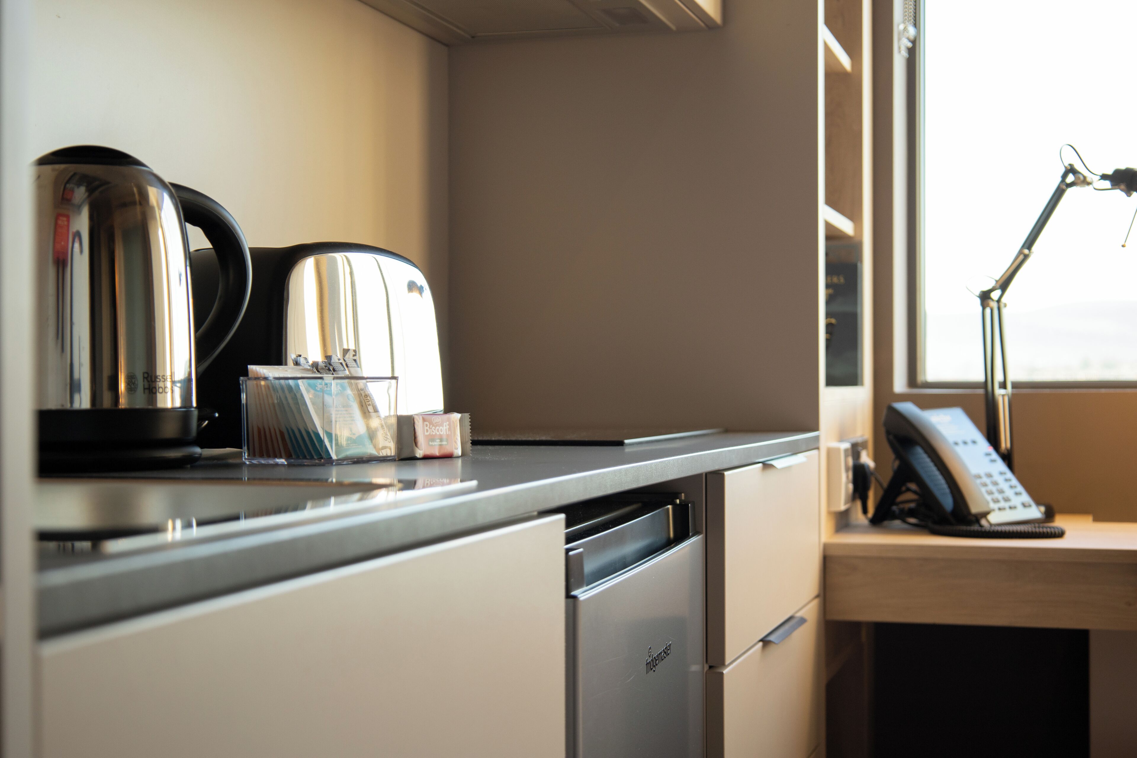 Een kitchenette met een koffiezetapparaat, broodrooster en een kleine koelkast. Er is een bureau met een telefoon en een lamp.