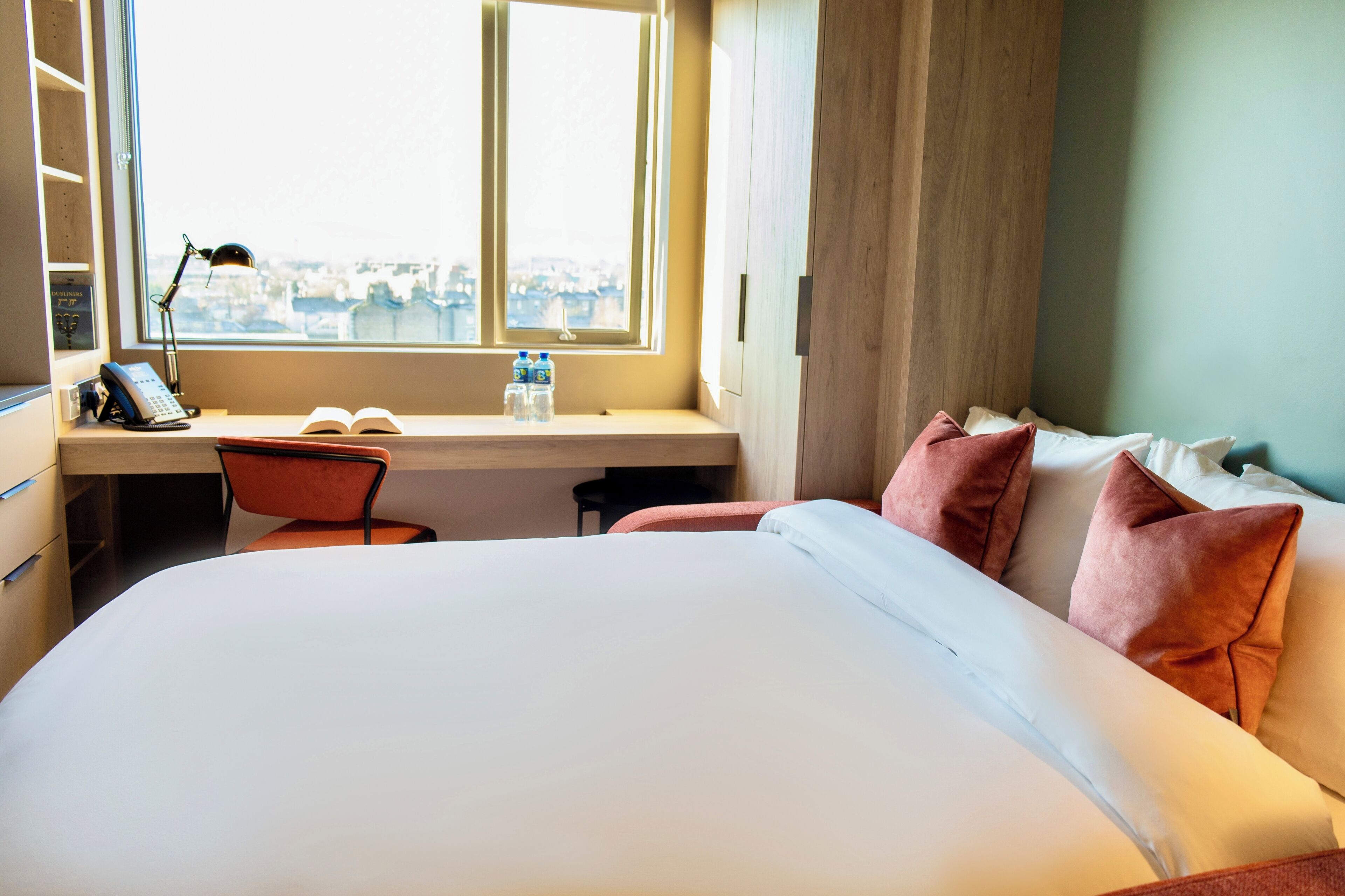 A hotel room with a bed, a desk with a chair, a window with a view, and a bookshelf.