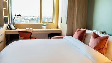 A hotel room with a bed, a desk with a chair, a window with a view, and a bookshelf.
