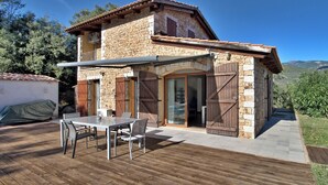 Une maison en pierre à deux étages, dotée d’une terrasse en bois et d’un espace extérieur aménagé et couvert.