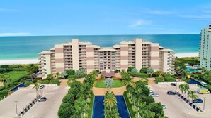 A multi-story hotel with a central courtyard, parking area, and a view of the beach. - Beachfront Condo With Views, Pools and Pickleball (Marco Island)