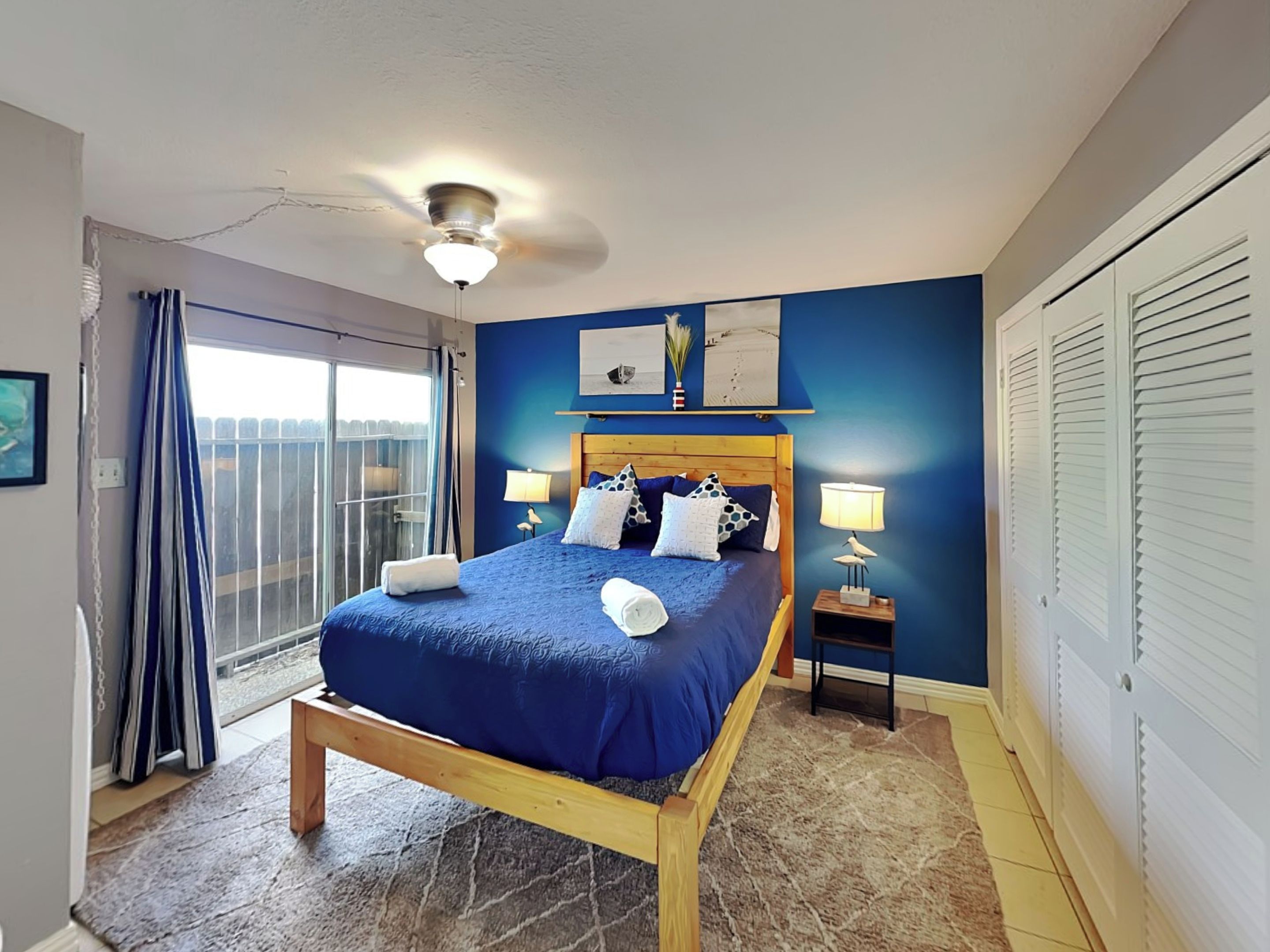 A bedroom with a bed, bedside lamps, a ceiling fan, a window with curtains, and a view of the outdoors.