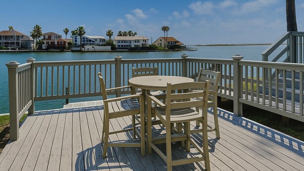 A wooden deck with a table and chairs overlooking a body of water with houses in the background. - Coastal Cozy and a View by Silver Sands Vacation Rentals (Corpus Christi)