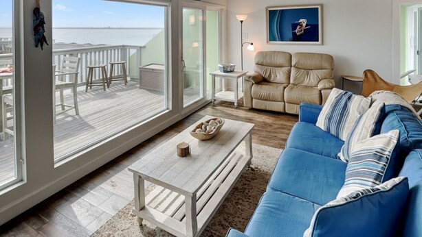 Ein geräumiges Wohnzimmer mit großem Fenster und Blick auf eine Terrasse, ein blaues Sofa, ein weißer Couchtisch und ein beiger Sessel.