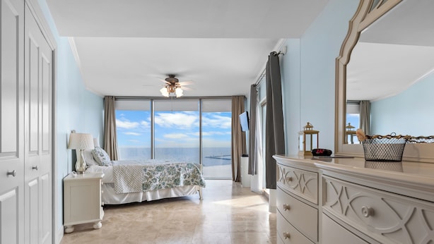 A bedroom with a bed, a nightstand, a dresser, a ceiling fan, and a large window with a view.