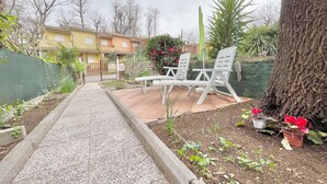 Un patio jardín con dos sillas blancas y una mesa, rodeado de plantas y un sendero que conduce a una puerta.