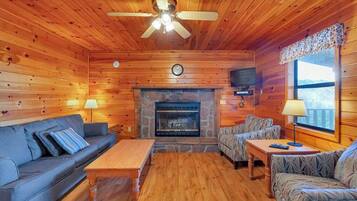 A log cabin interior with a fireplace, wooden walls, a ceiling fan, and a sofa.
