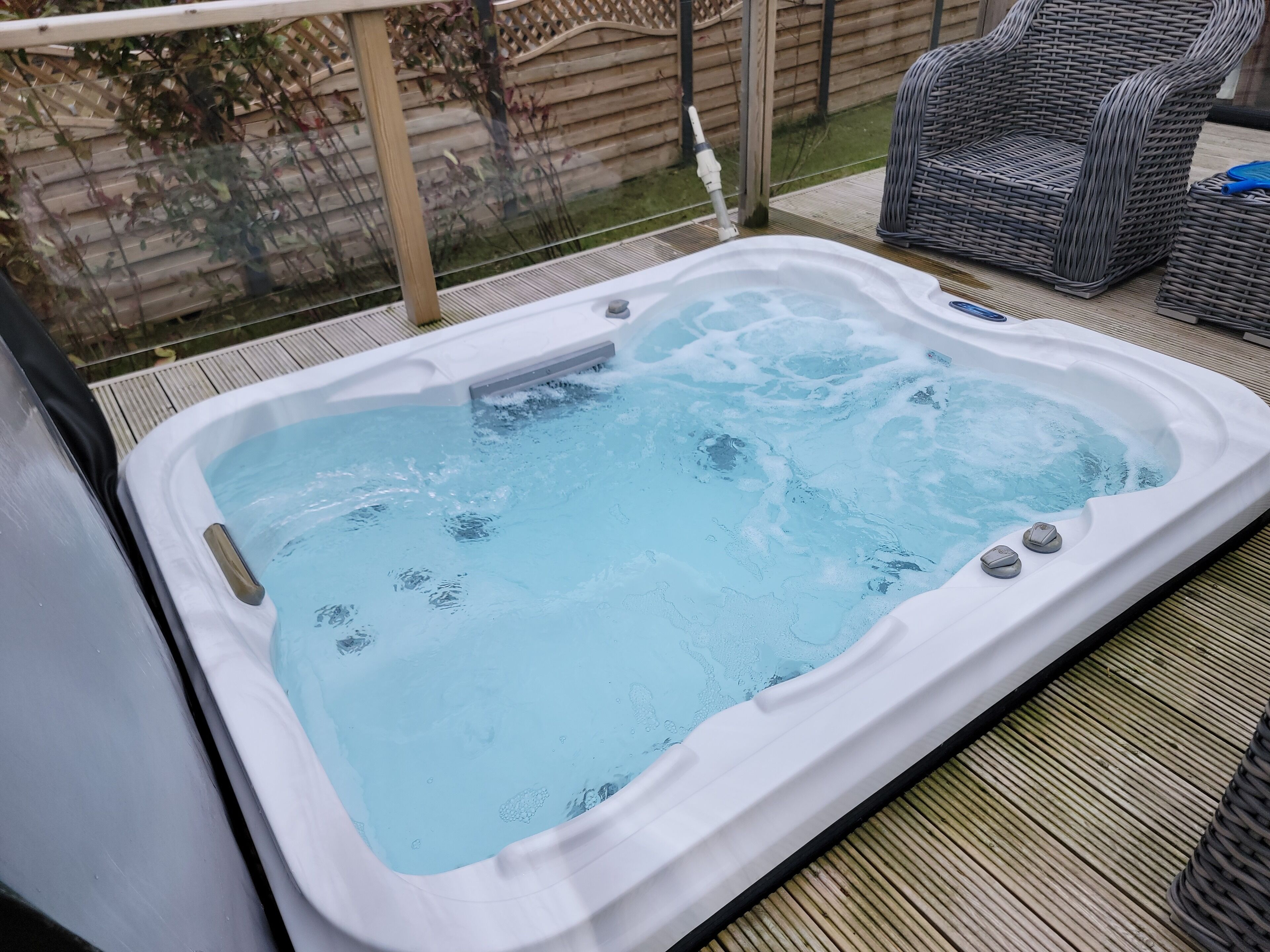A hot tub on a wooden deck with a wicker chair and a fence in the background.