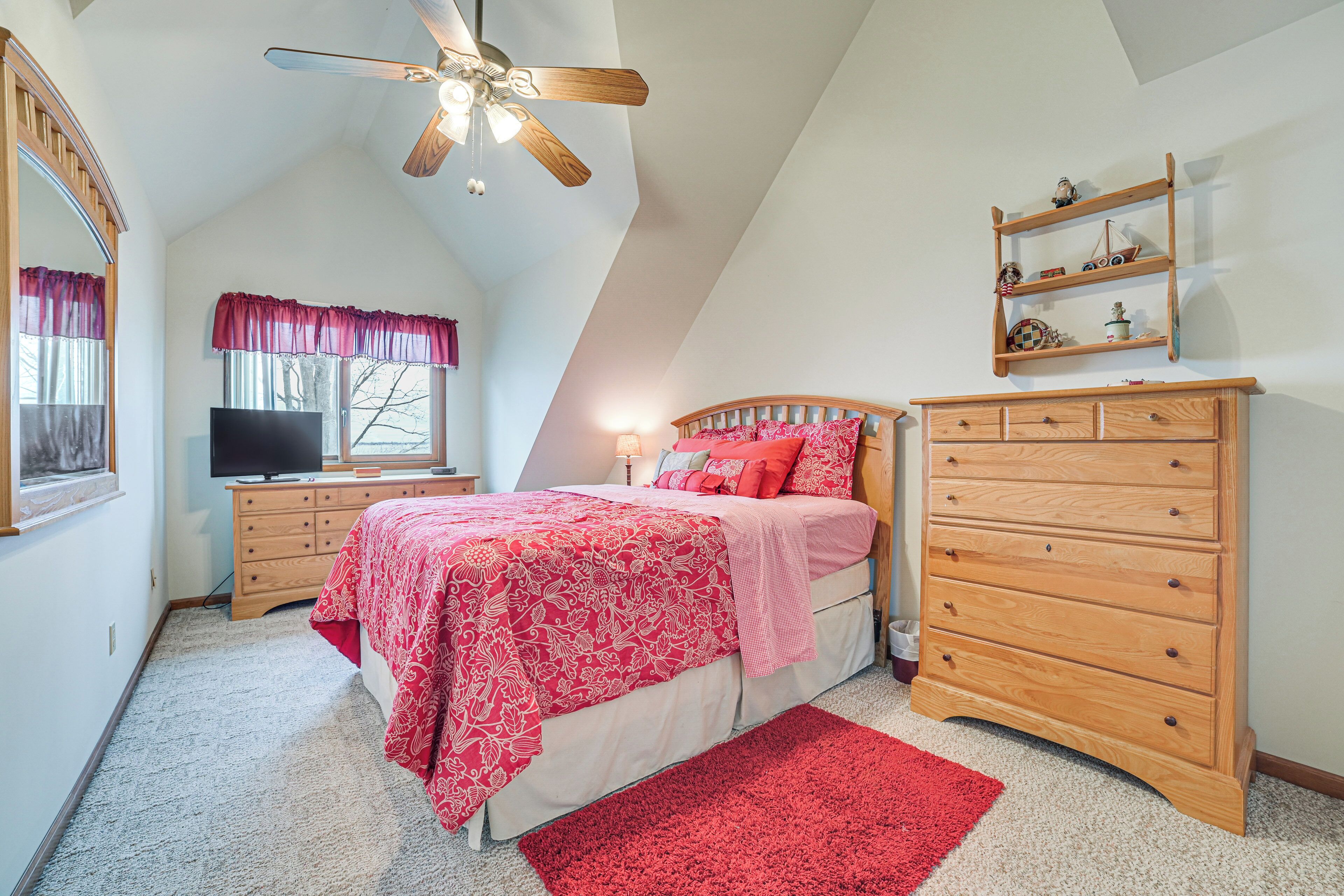 A bedroom with a sloped ceiling, a bed with a red and white patterned bedspread, a wooden dresser, a nightstand, a television, and a window with a red valance.