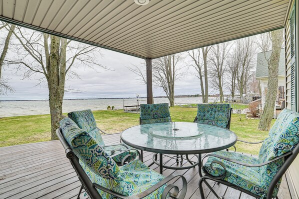 Een overdekt terras met een glazen tafel en gestreept meubilair, met uitzicht op een gazon en een waterpartij.