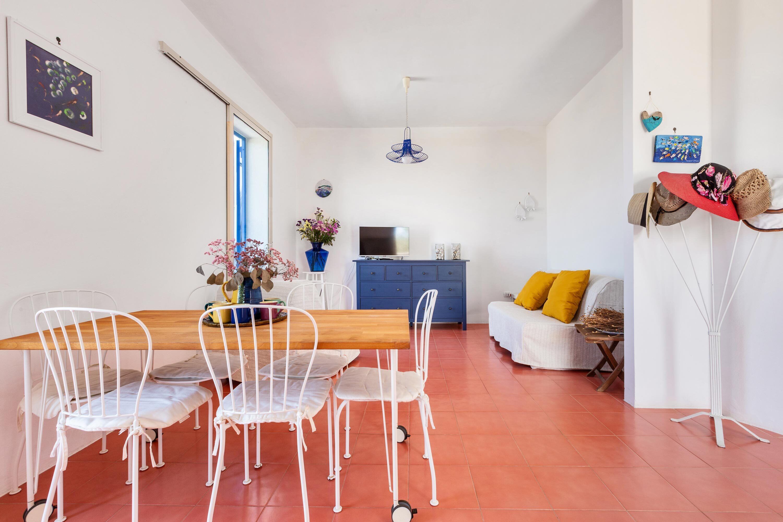 A dining area with a wooden table and white chairs, a blue cabinet, a TV, and a sofa with yellow cushions.