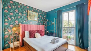 Una camera da letto con una parete color petrolio, carta da parati a motivi floreali, una testiera imbottita rosa, pavimento in legno, un comodino con una lampada e una finestra con tende.